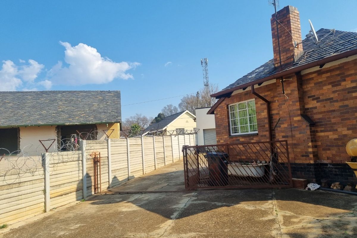 Exterior view of the property featuring a brick facade, chimney, concrete driveway, and perimeter wall with security fencing.