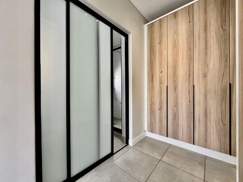 A modern bedroom area featuring sleek wood-finish built-in cupboards, large format floor tiles, and a contemporary black-framed sliding glass door.