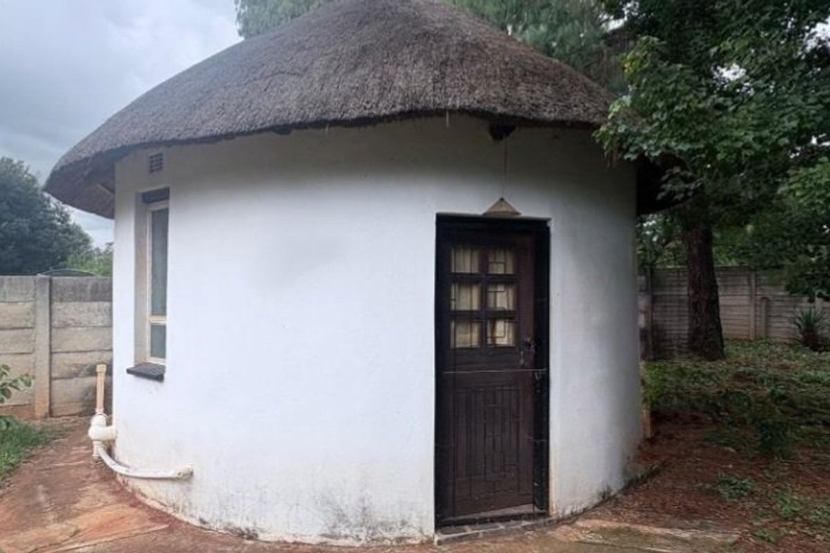 A detached circular outbuilding featuring a traditional thatched roof, timber door, and casement window, situated within a garden setting with concrete boundary walls.