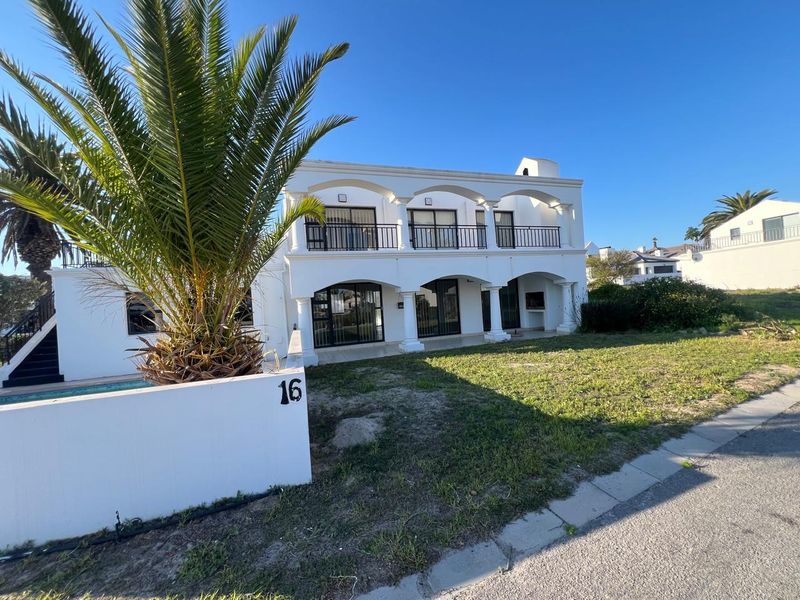 A two-story white house featuring arched balconies, a swimming pool, and a built-in braai area, set against a clear blue sky.