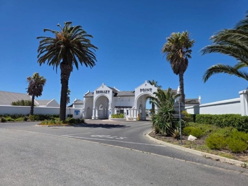The main entrance gate to the Shelley Point estate, featuring a white architectural archway, paved access road, and established palm tree landscaping.