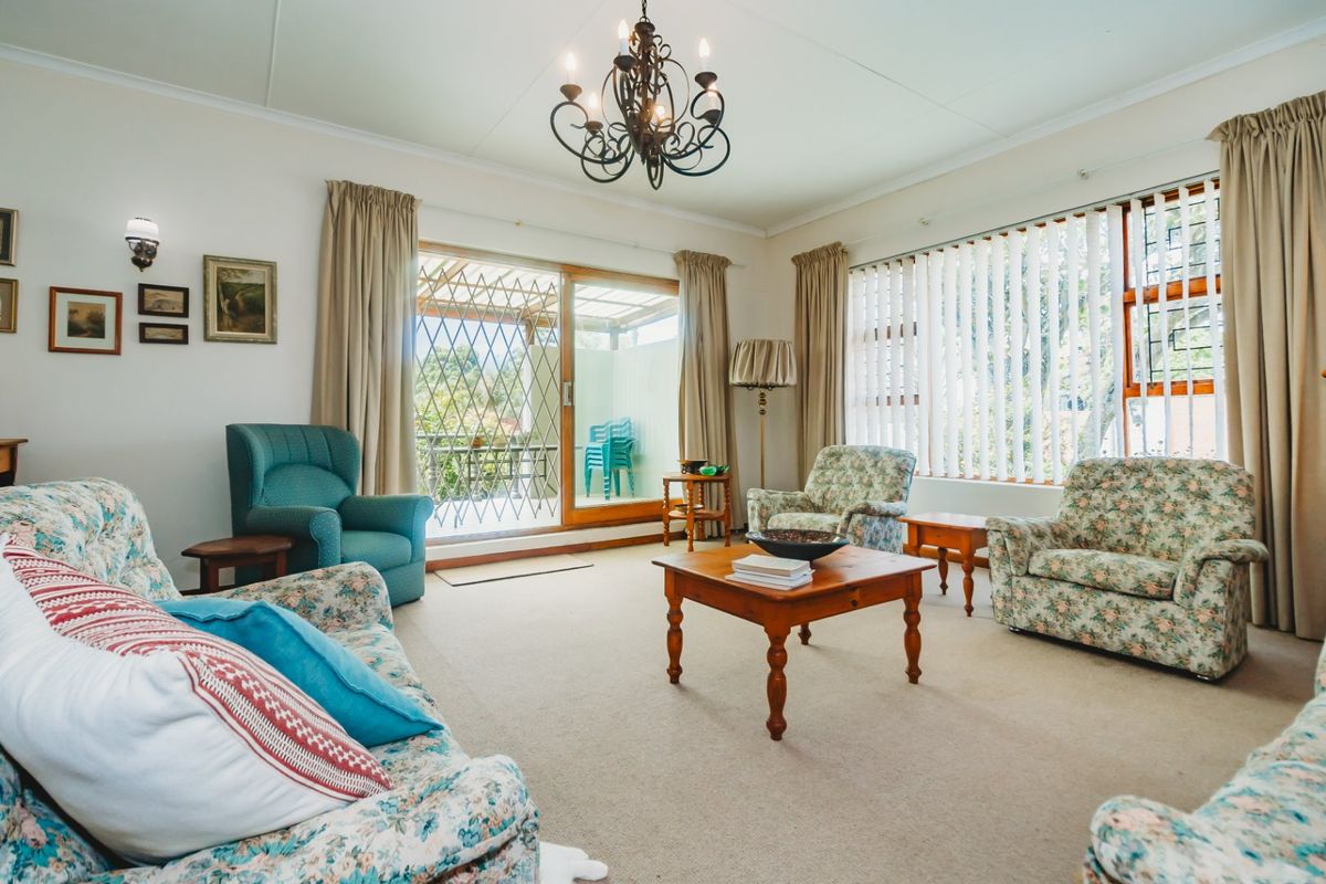 A spacious living room featuring carpeted flooring, a chandelier, and large windows with vertical blinds, providing access to an outdoor patio area.