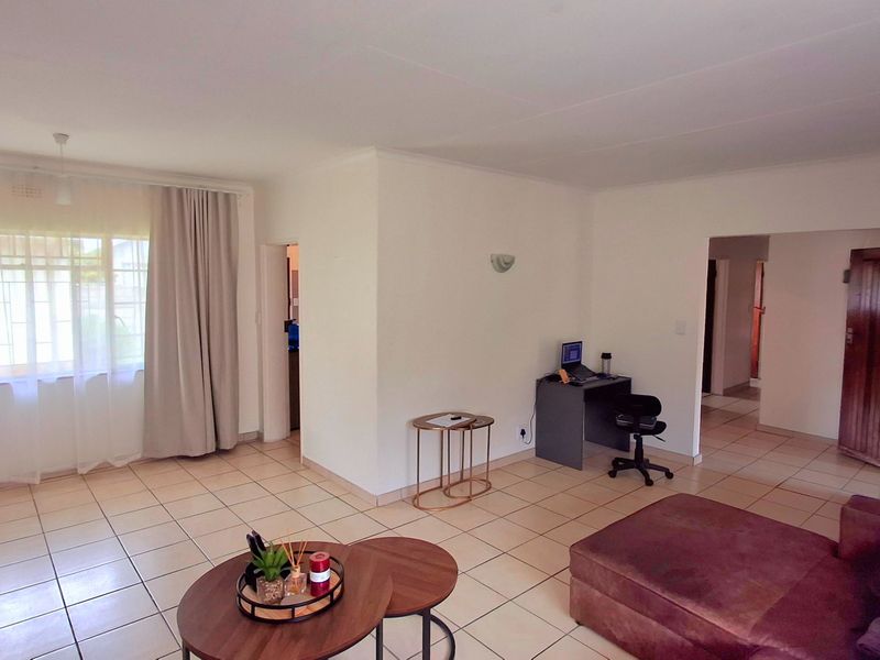 A spacious living area featuring neutral tiled flooring, large windows with security bars, and a functional layout connecting to adjacent rooms.