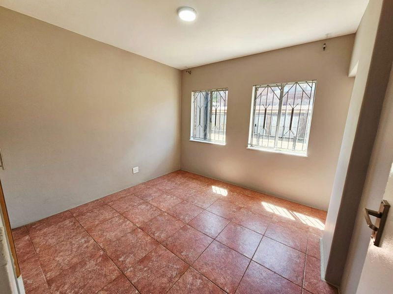 A clean, empty bedroom featuring tiled flooring, neutral wall paint, and windows fitted with security bars.