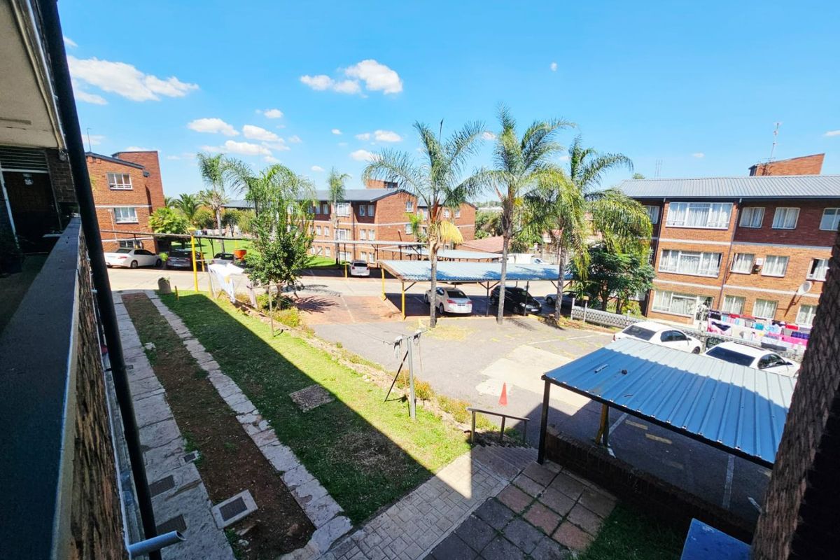 View of the apartment complex exterior, featuring paved parking areas, covered carports, and communal garden spaces between the brick residential buildings.