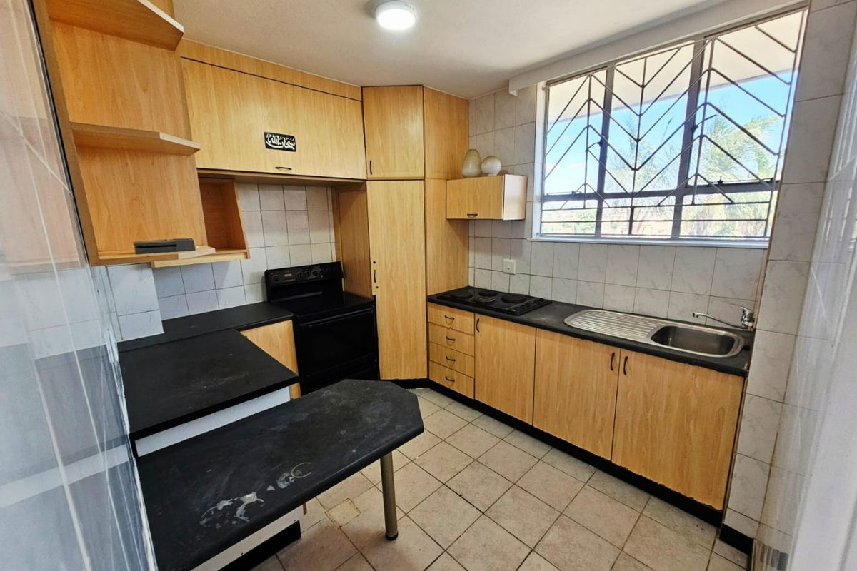 A functional kitchen featuring light wood-finish cabinetry, black countertops, a built-in oven and hob, and tiled flooring with a window providing natural light.