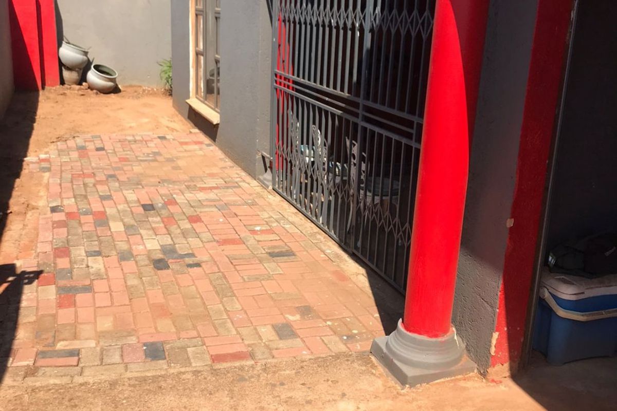 Rear exterior area featuring brick paving, a security gate, and a decorative column, with an adjacent unfinished soil ground area.