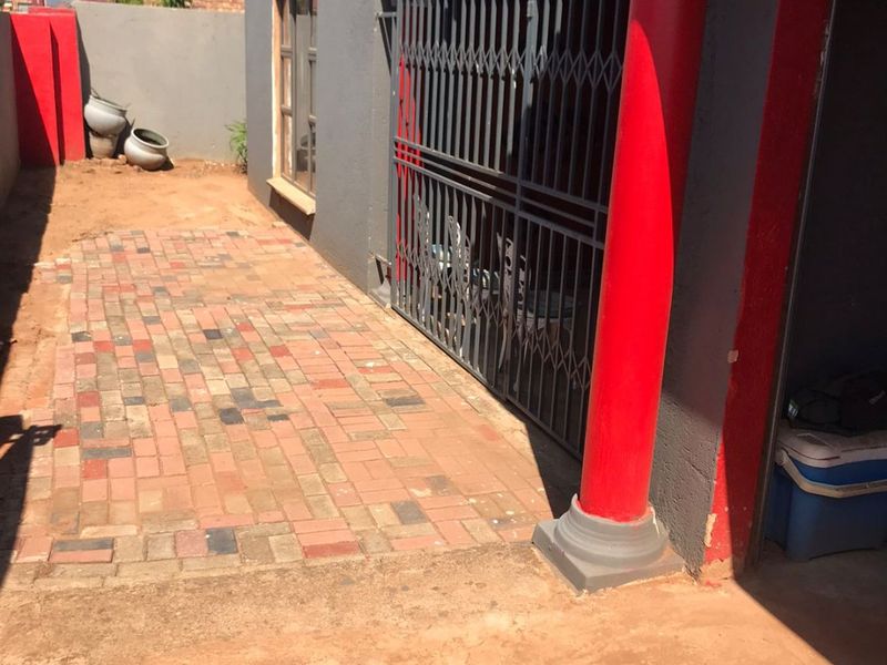 Rear exterior area featuring brick paving, a security gate, and a decorative column, with an adjacent unfinished soil ground area.