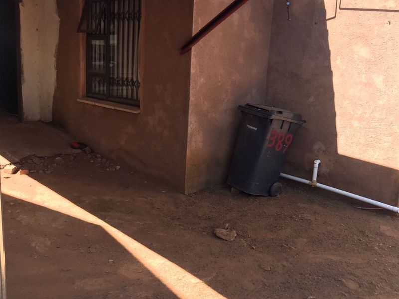 An outdoor covered patio area featuring a metal carport roof, security gate, and unfinished exterior wall plaster with unpaved ground.