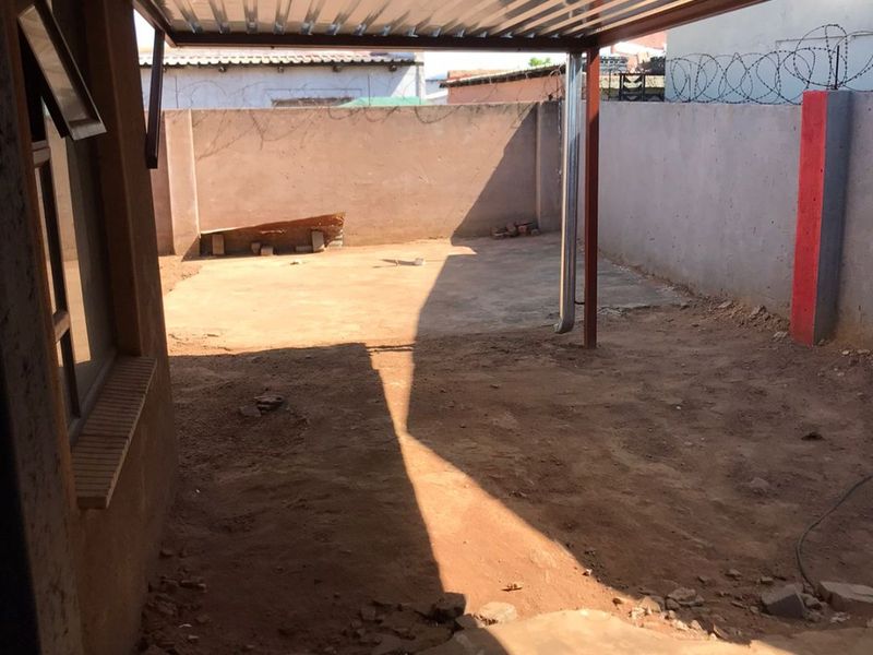 A covered outdoor patio area featuring a corrugated metal roof, concrete flooring, and a perimeter boundary wall with security fencing.