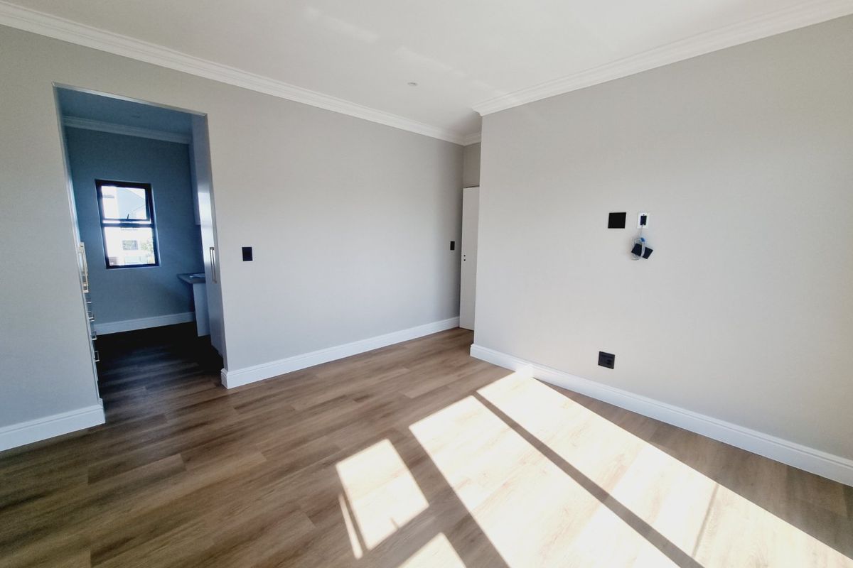 A bright, empty living area featuring modern laminate flooring, neutral walls, and coving, with an adjacent view into a secondary utility or kitchen space.