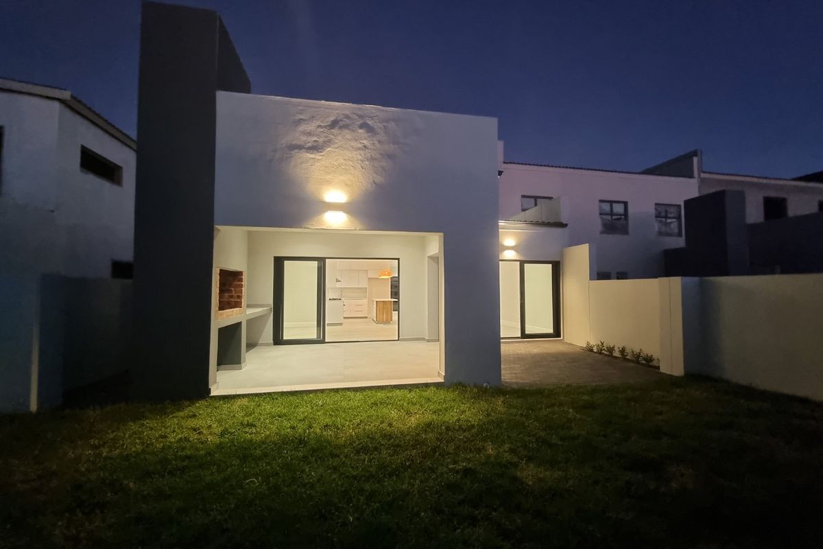 Modern rear exterior view featuring a covered patio with a built-in braai, sliding glass doors, and a well-maintained garden at dusk.