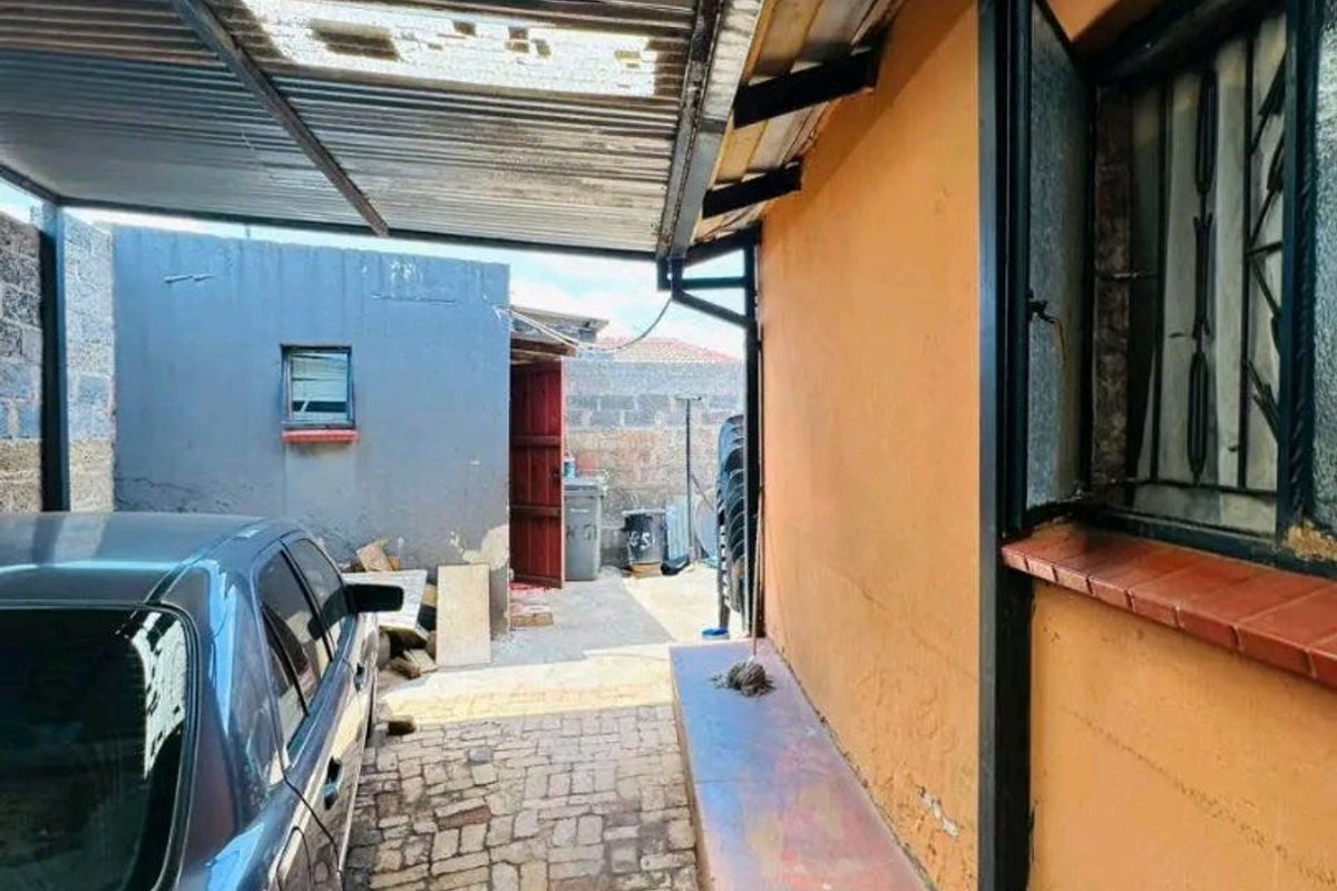 A covered outdoor area featuring a corrugated metal roof, brick paving, and a concrete ledge along the exterior wall of the house.