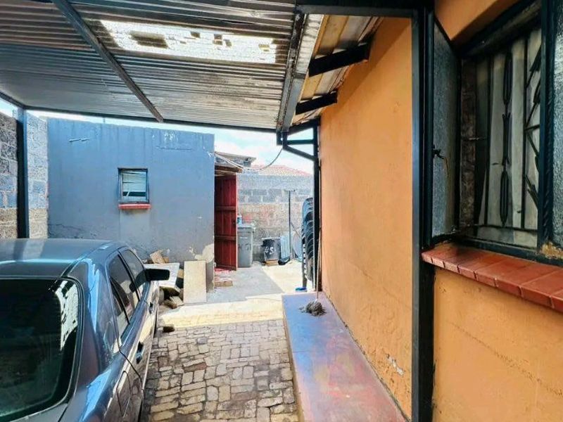 A covered outdoor area featuring a corrugated metal roof, brick paving, and a concrete ledge along the exterior wall of the house.