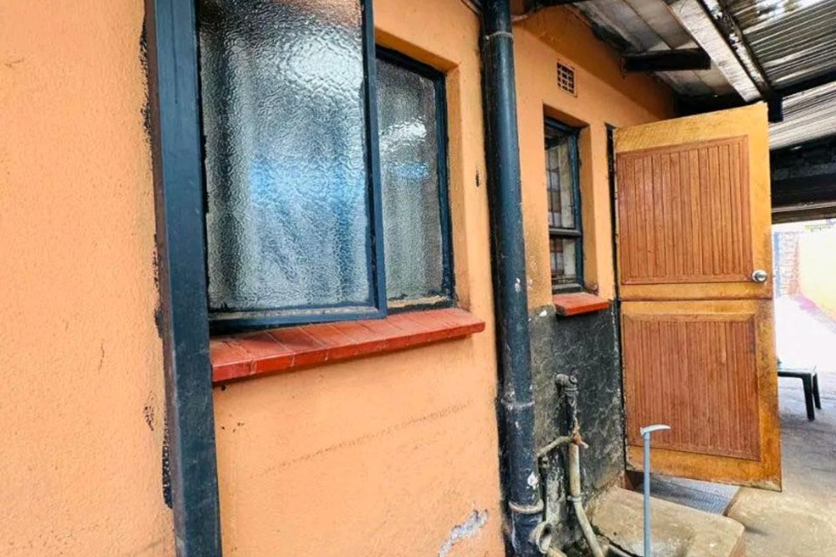 Rear exterior view of the property showing weathered masonry, steel window frames, and a timber door under a corrugated metal roof structure.