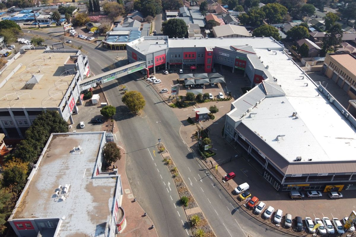Aerial view of a commercial complex featuring multiple buildings, a connecting pedestrian bridge, paved parking areas with shade structures, and surrounding street access.