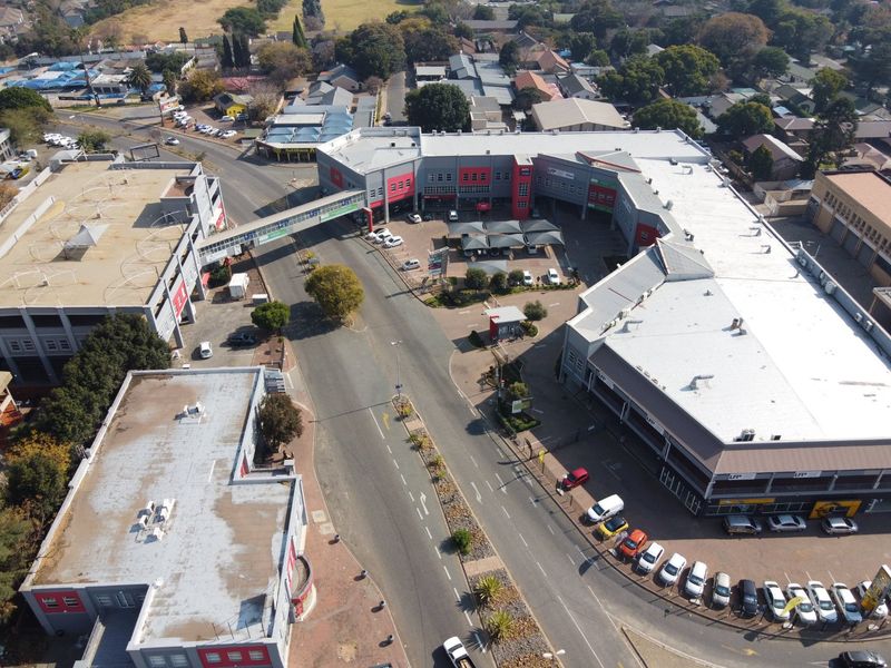 Aerial view of a commercial complex featuring multiple buildings, a connecting pedestrian bridge, paved parking areas with shade structures, and surrounding street access.