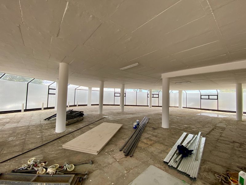 A large, open-plan commercial space in shell condition, featuring structural columns, a sloped glass facade, and unfinished concrete flooring.