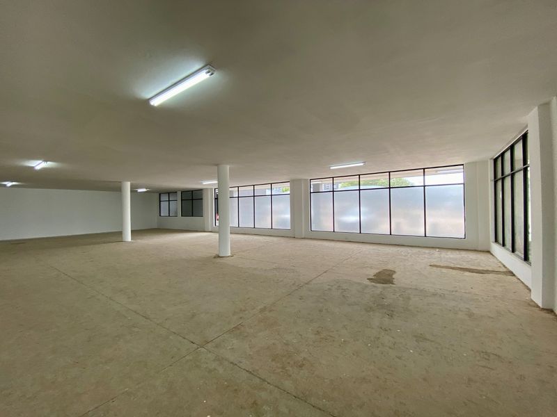A spacious, open-plan commercial interior featuring structural columns, large windows for natural light, and unfinished concrete flooring.