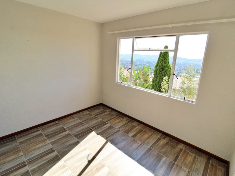 A bright, empty bedroom featuring modern wood-look tiled flooring, neutral walls, and a large window offering scenic views.