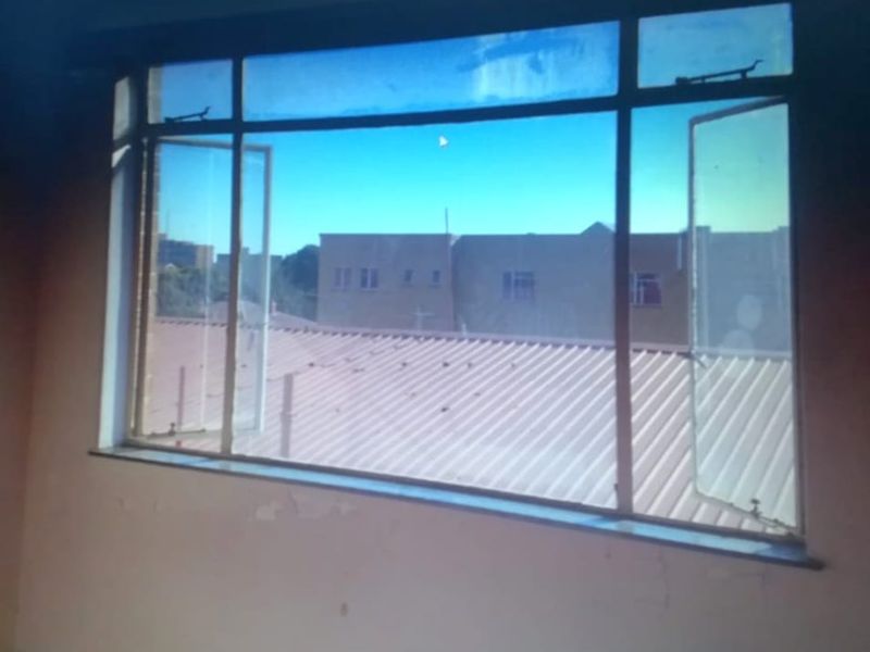 An empty bedroom featuring a large metal-framed window with views of the surrounding neighborhood and rooflines, requiring cosmetic wall repairs.