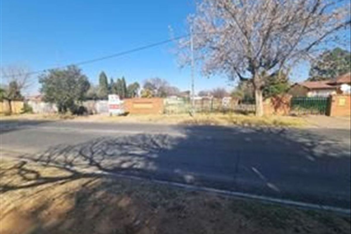 Street view of a vacant residential plot featuring mature trees and a boundary wall, situated along a paved road.