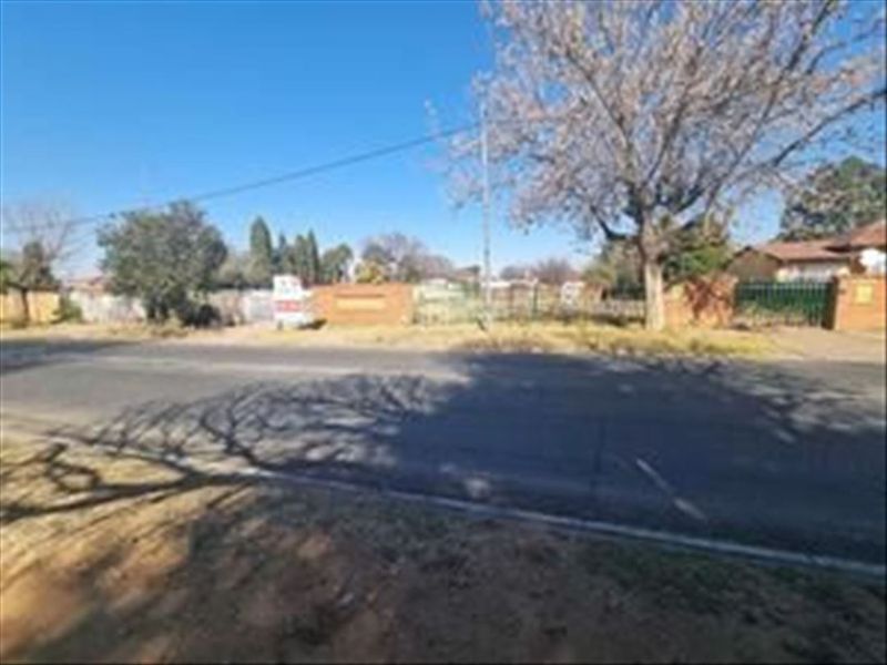 Street view of a vacant residential plot featuring mature trees and a boundary wall, situated along a paved road.