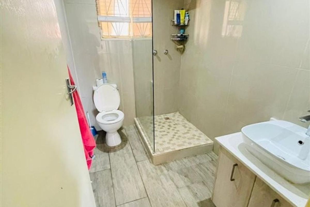 A functional bathroom featuring a glass-enclosed shower, vessel sink vanity, and neutral floor tiling with security bars on the window.