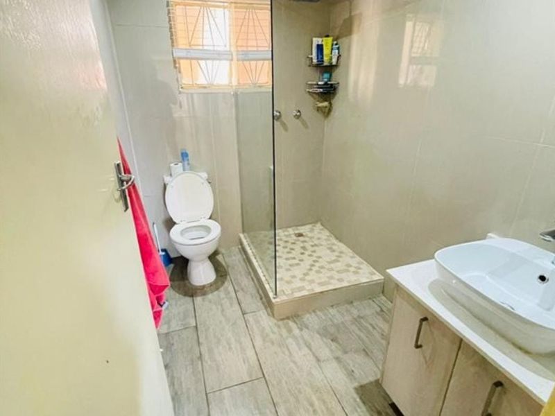 A functional bathroom featuring a glass-enclosed shower, vessel sink vanity, and neutral floor tiling with security bars on the window.
