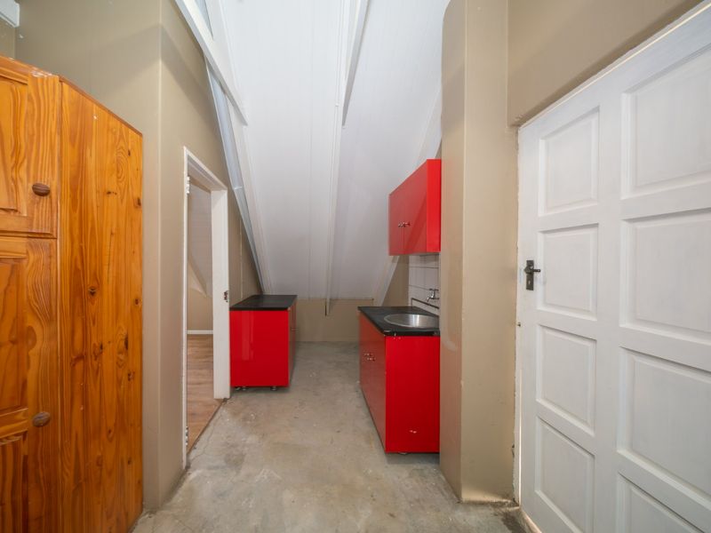 A compact scullery or utility area featuring red built-in cabinetry, a stainless steel sink, and a sloped ceiling with unfinished concrete flooring.