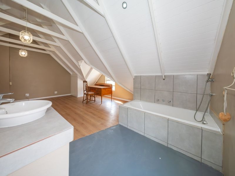 An open-plan attic bathroom featuring a built-in bathtub with grey tiling, a basin, timber flooring, and exposed white-painted rafters.