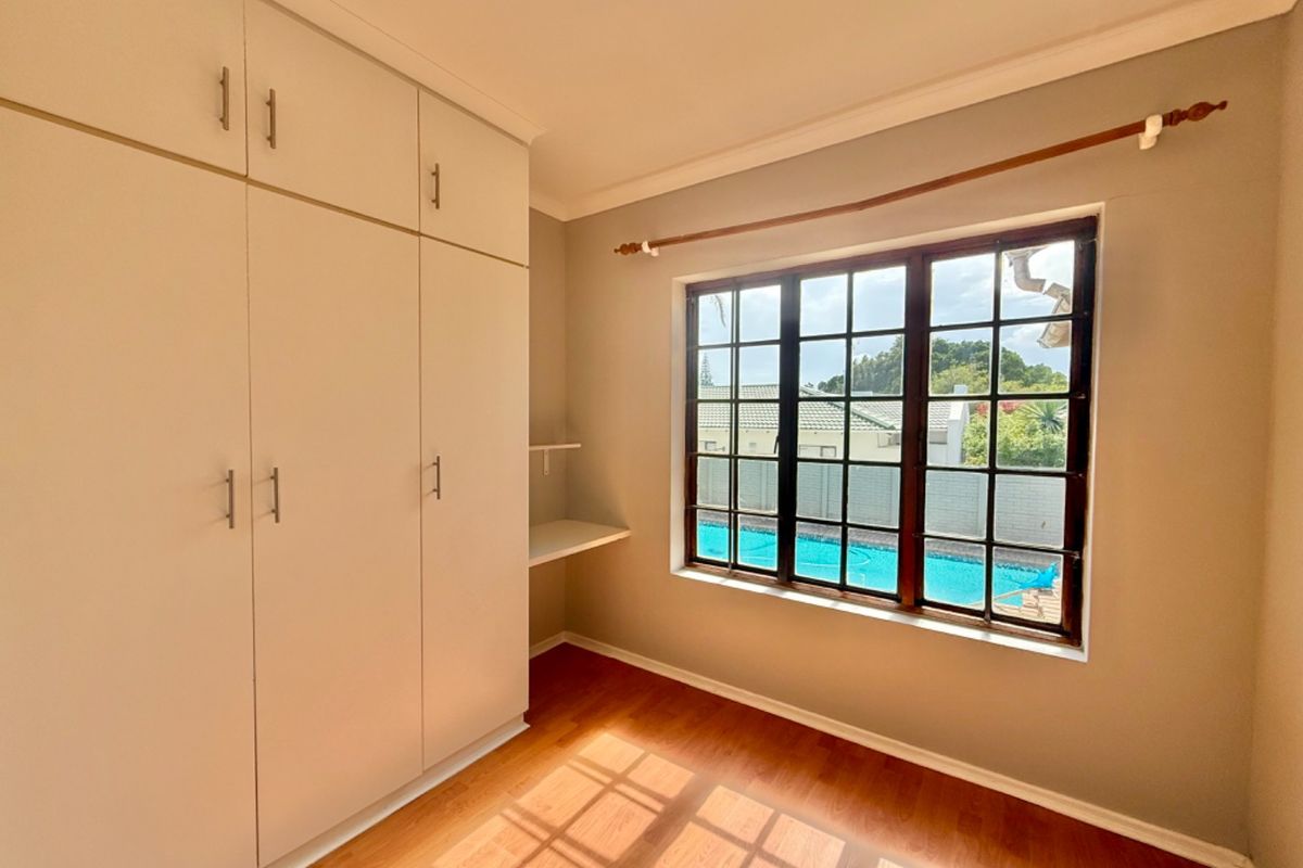 A bright, empty bedroom featuring laminate flooring, built-in cupboards, a built-in desk shelf, and a window overlooking the swimming pool area.