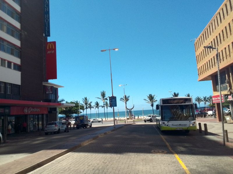 A street-level view of a coastal urban area featuring commercial buildings, a public bus, and a clear view towards the beach.