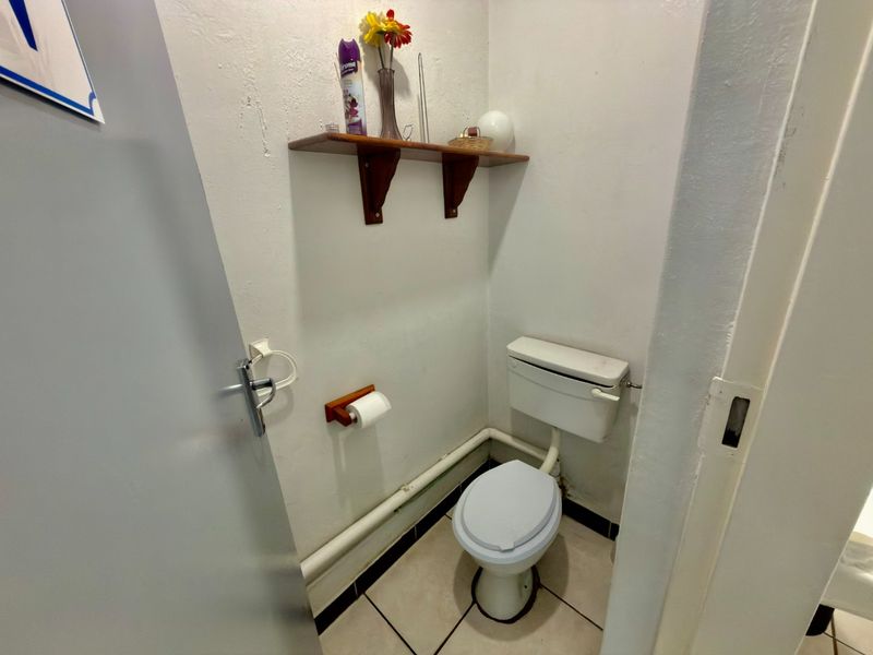 A compact commercial bathroom featuring a standard toilet, wall-mounted shelving, and exposed plumbing fixtures with tiled flooring.