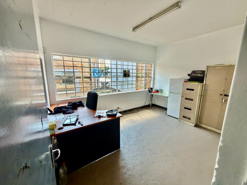 A functional office space featuring a large window with security bars, fluorescent lighting, and basic flooring, suitable for commercial use.