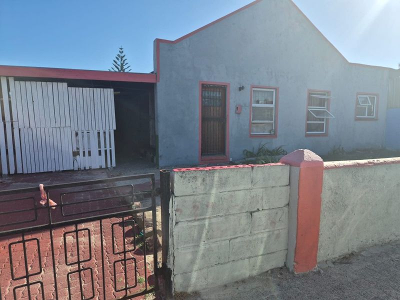 Front view of a single-story house featuring a concrete block boundary wall, metal security gate, paved driveway, and an attached carport.