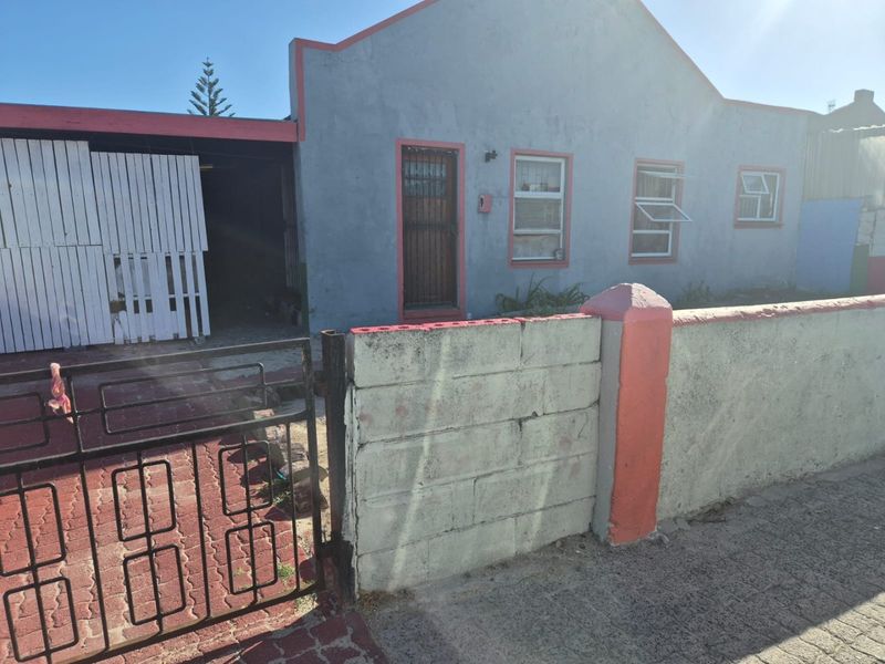Front elevation of a single-story house featuring a concrete block boundary wall, metal security gate, paved driveway, and a covered carport area.