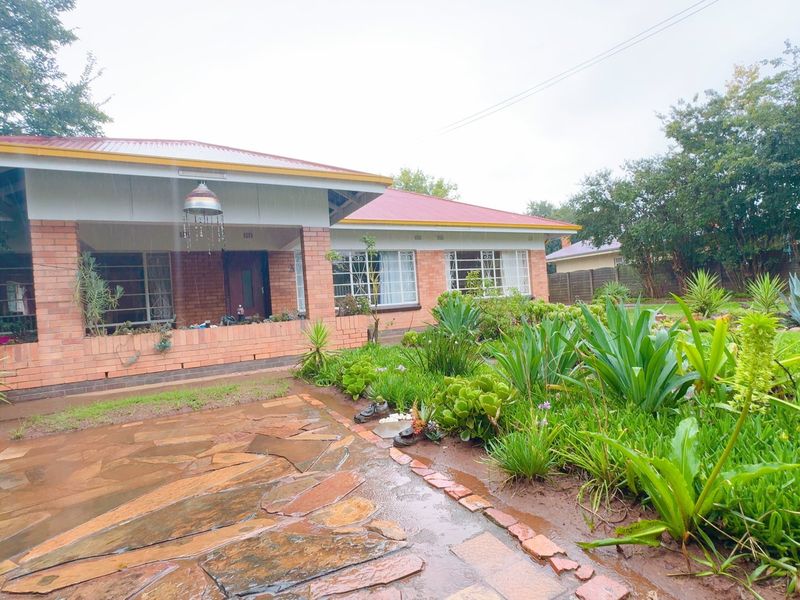 A charming single-story brick house featuring a covered front patio, metal roof, and established garden with stone-paved walkway.
