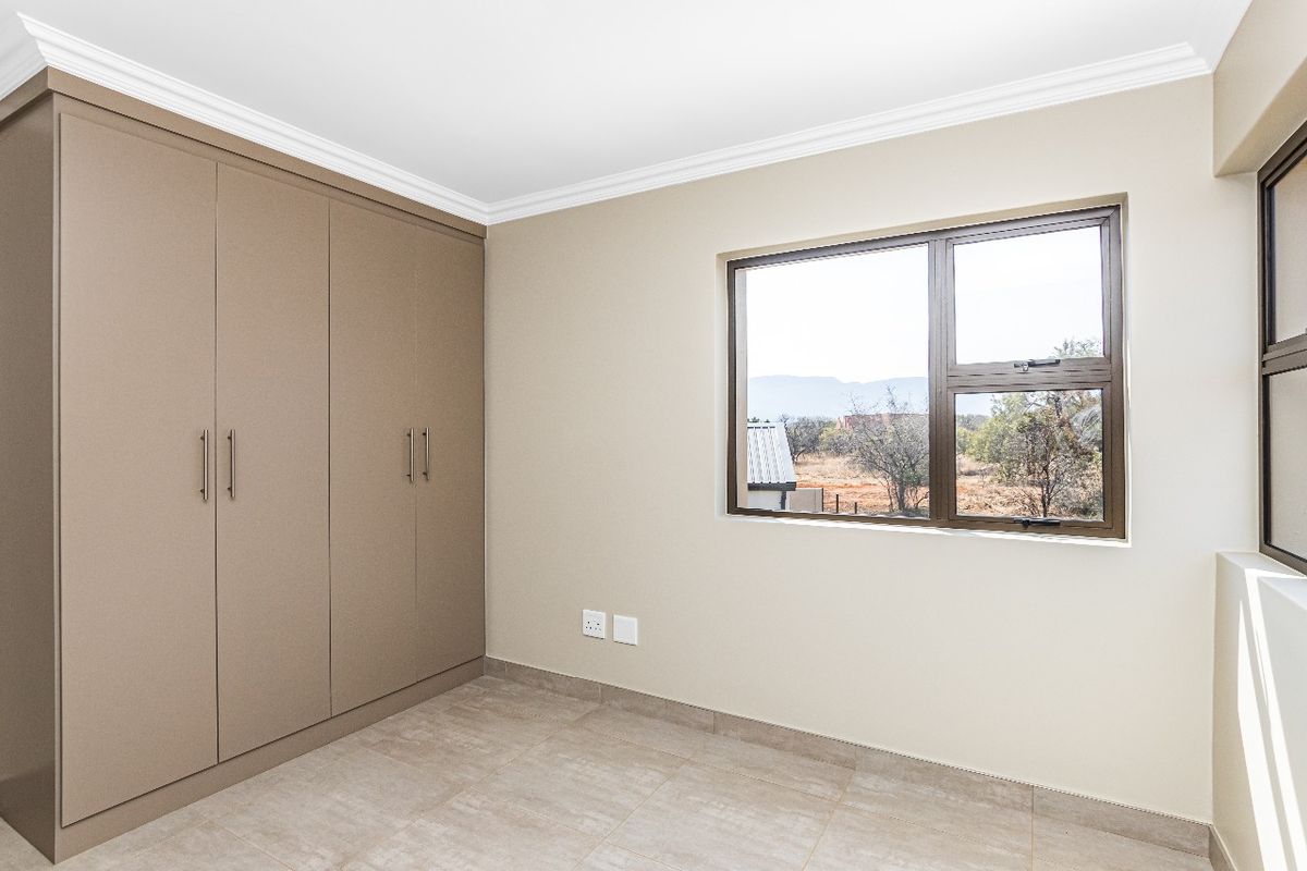 A clean, empty bedroom featuring neutral tiled flooring, built-in cupboards, and aluminium window frames with a view of the surrounding landscape.
