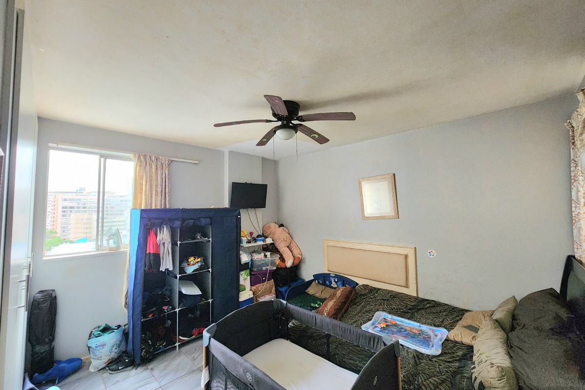 A bedroom featuring tiled flooring, a ceiling fan, and a sliding window. The room shows signs of wear including ceiling staining and peeling paint.