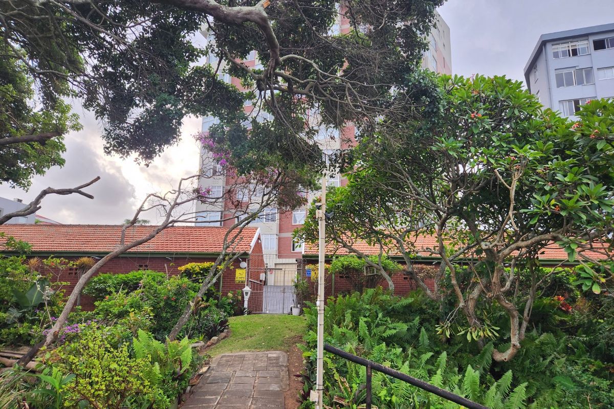 A view of the rear garden area featuring a paved walkway, established landscaping, and a brick boundary wall with a security gate.
