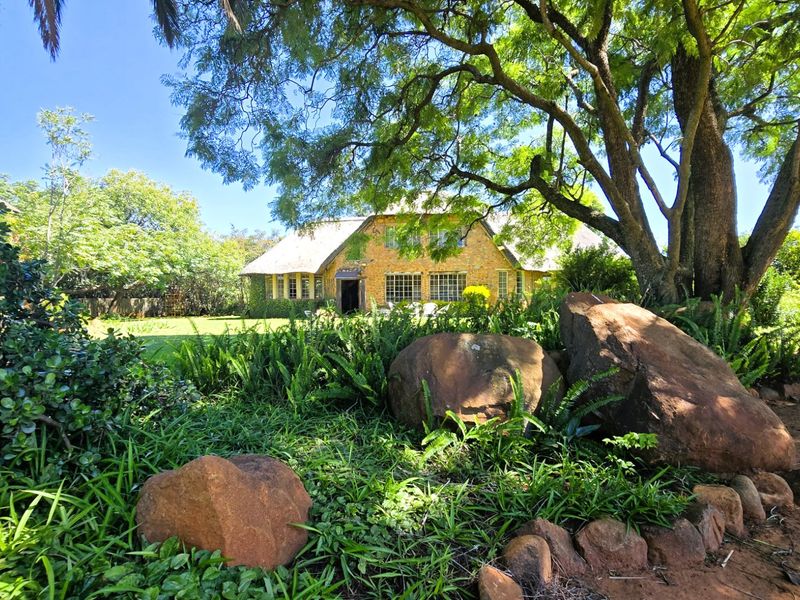 A charming farmhouse featuring traditional stone cladding and a thatched roof, set within a lush, mature garden with large feature rocks.