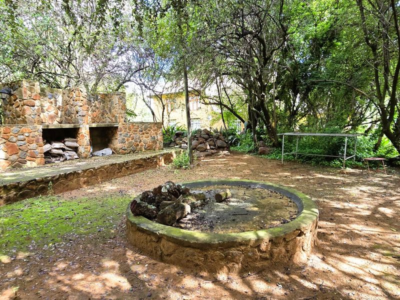 A rustic outdoor garden area featuring a stone-built braai and a circular stone fire pit, set within a mature, wooded landscape.