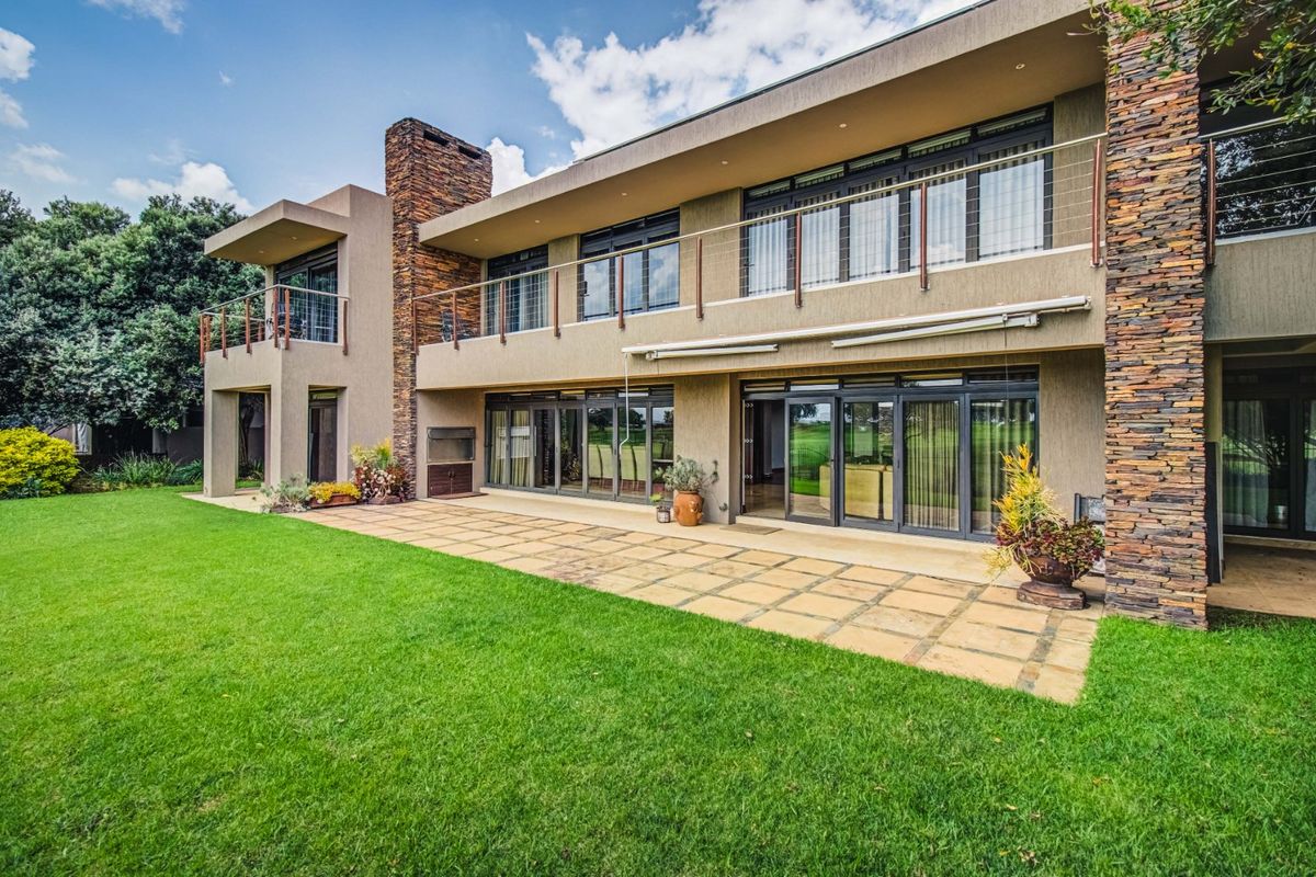 Spacious rear exterior featuring a paved patio, built-in braai, stone-clad pillars, and a large balcony overlooking a well-maintained garden.