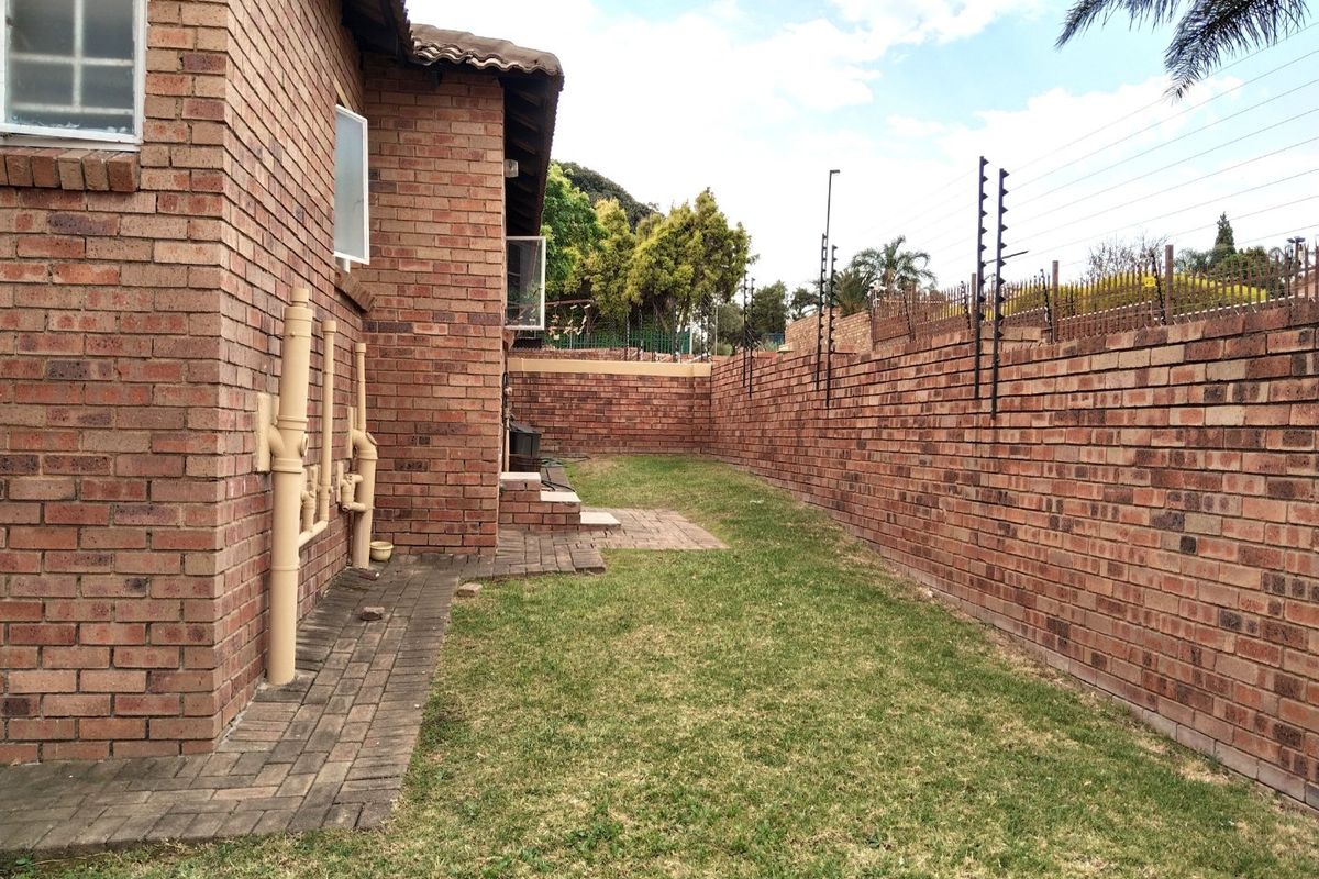 A side view of the townhouse exterior featuring a brick facade, paved walkway, small lawn area, and secure boundary wall with electric fencing.