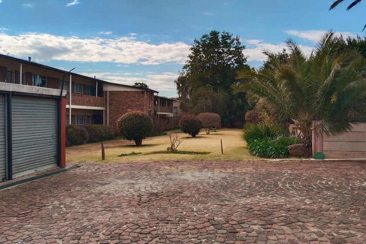 Exterior view of an apartment complex featuring a brick facade, paved driveway, garage units, and a shared garden area.