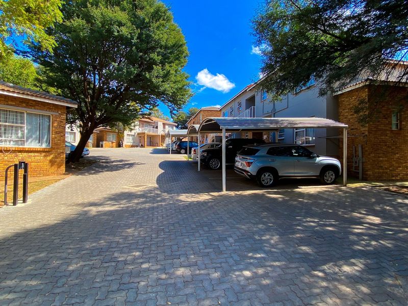 Exterior view of an apartment complex featuring a paved driveway, covered parking carports, and established landscaping with mature trees.
