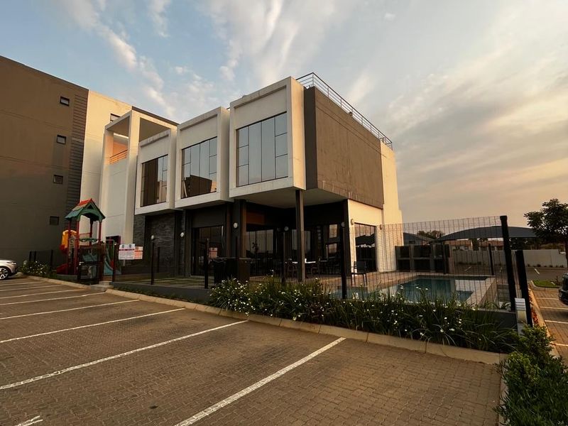 Modern apartment complex exterior featuring a paved parking area, swimming pool, and contemporary architectural design with large glass windows.