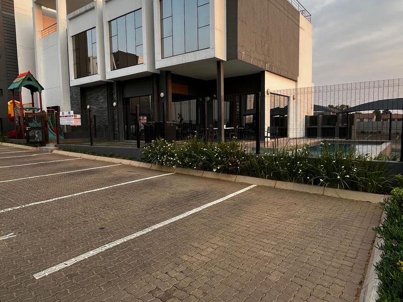 Modern apartment complex exterior featuring a paved parking area, swimming pool, and contemporary architectural design with large glass windows.