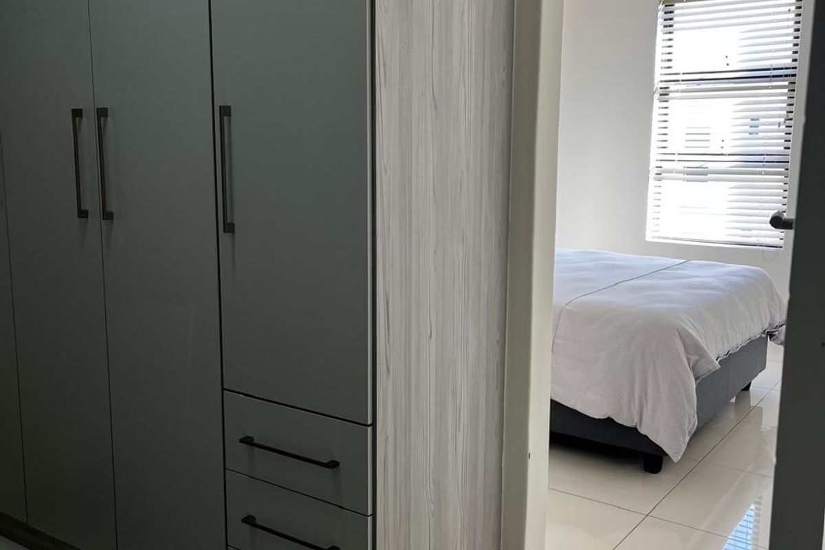 A bedroom featuring modern built-in cupboards with a light wood and grey finish, polished floor tiles, and a window with venetian blinds.